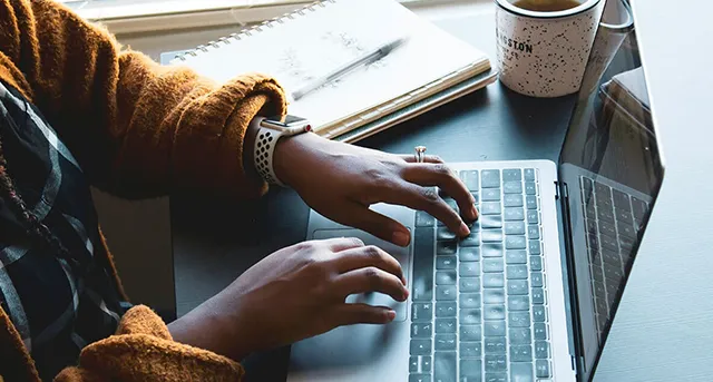 Hands typing on a laptop with coffee and a notepad on the desk nearby