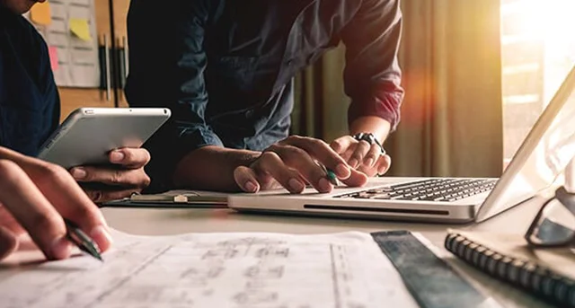 Hands of 2 people working, holding a tablet and using a laptop.